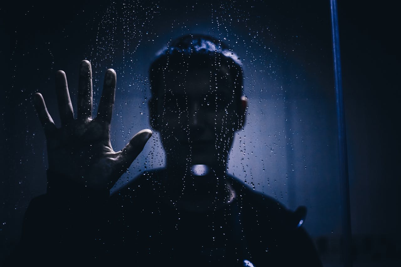 Silhouette of a person with hand against a rain-covered glass, evoking mystery and introspection.