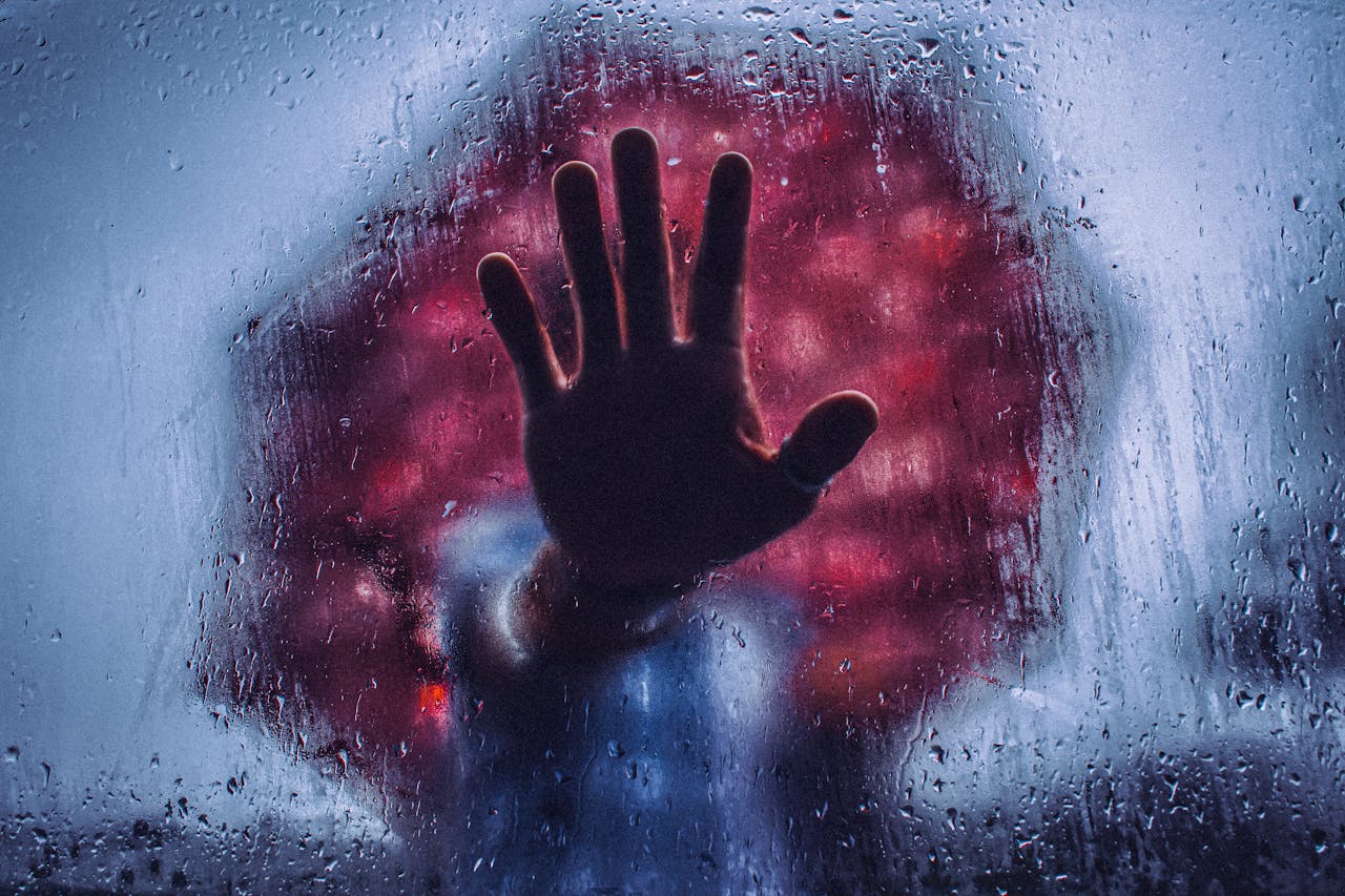 A hand pressed against wet glass with raindrops, creating a dramatic atmosphere.